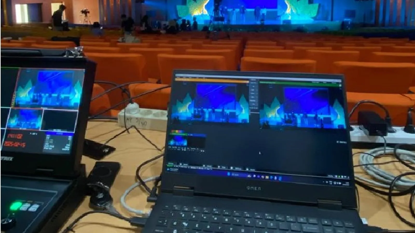 Meja operator dengan switcher video dan laptop streaming siap dipakai di sebuah auditorium.